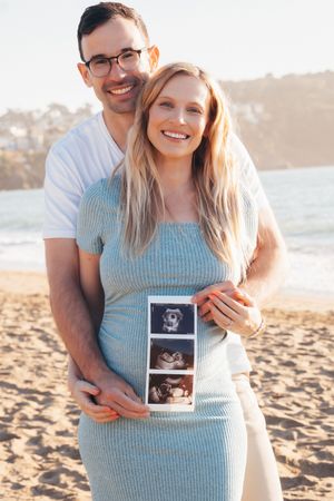 Smiling pregnant couple on beach holding ultrasound photos, man embracing woman in blue dress with ocean backdrop.