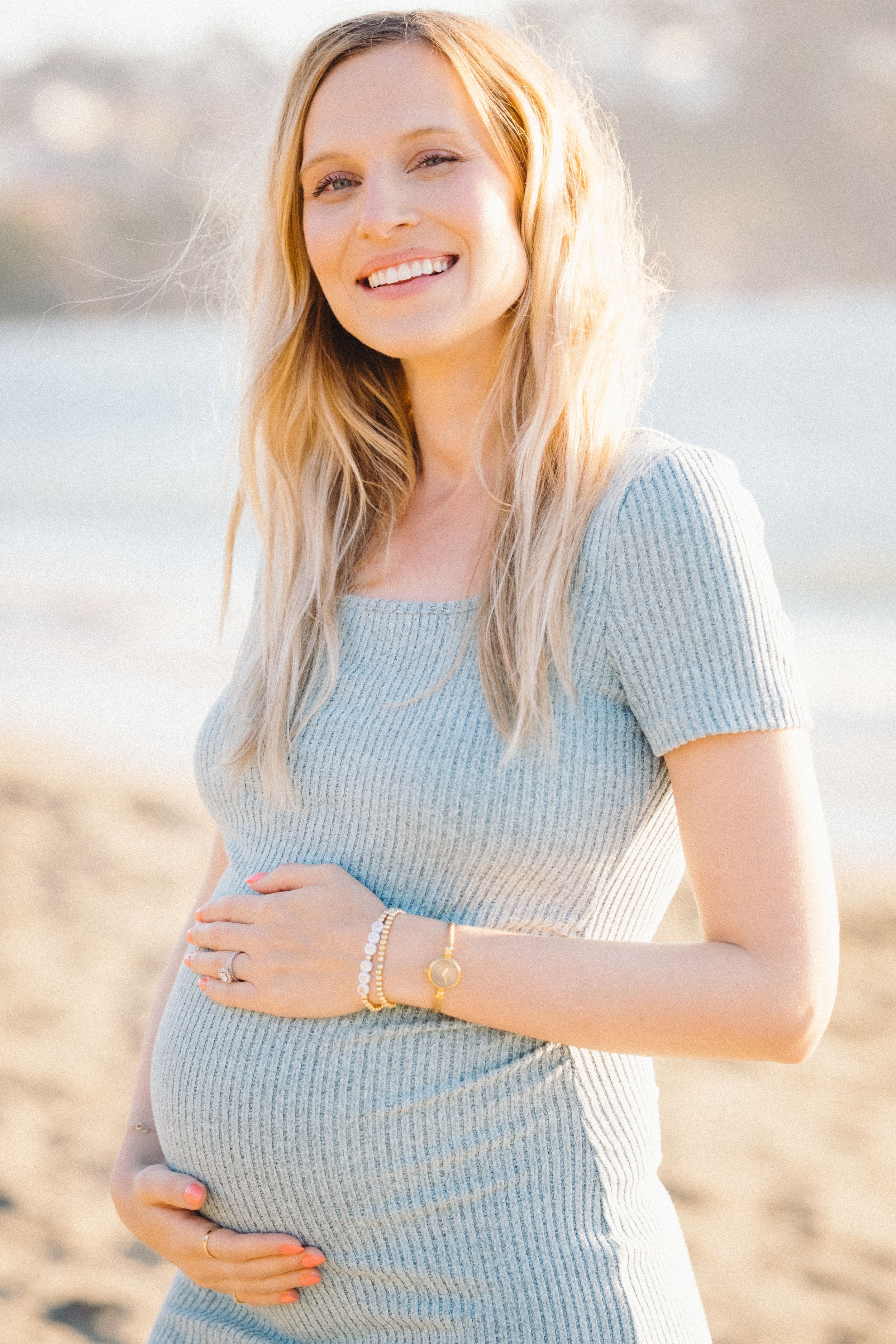 Smiling pregnant woman in a light blue ribbed dress cradling her baby bump on a sunny beach.