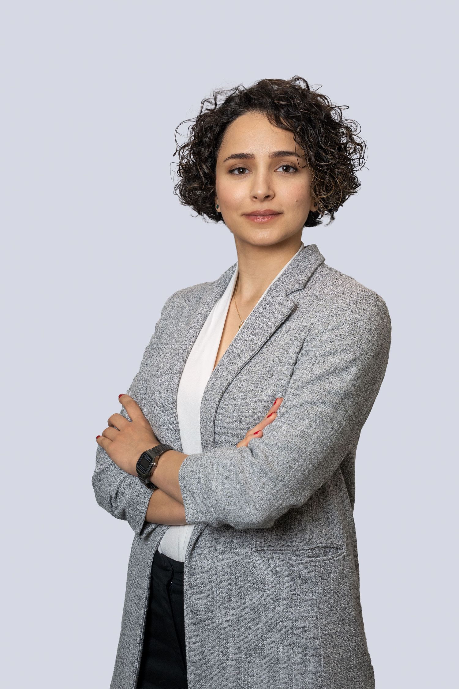 Professional wearing a gray blazer and white top poses against light background with curly hair and confident stance.
