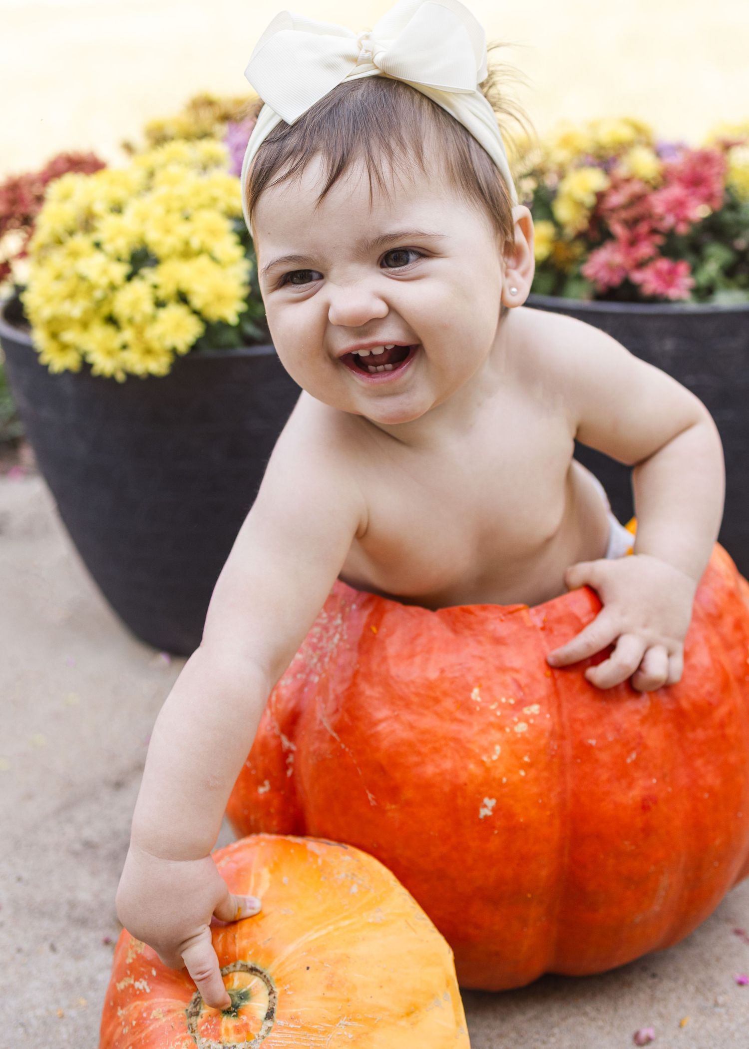 A baby in a white shirt sits on a bright orange pumpkin while surrounded by yellow and pink flowers.