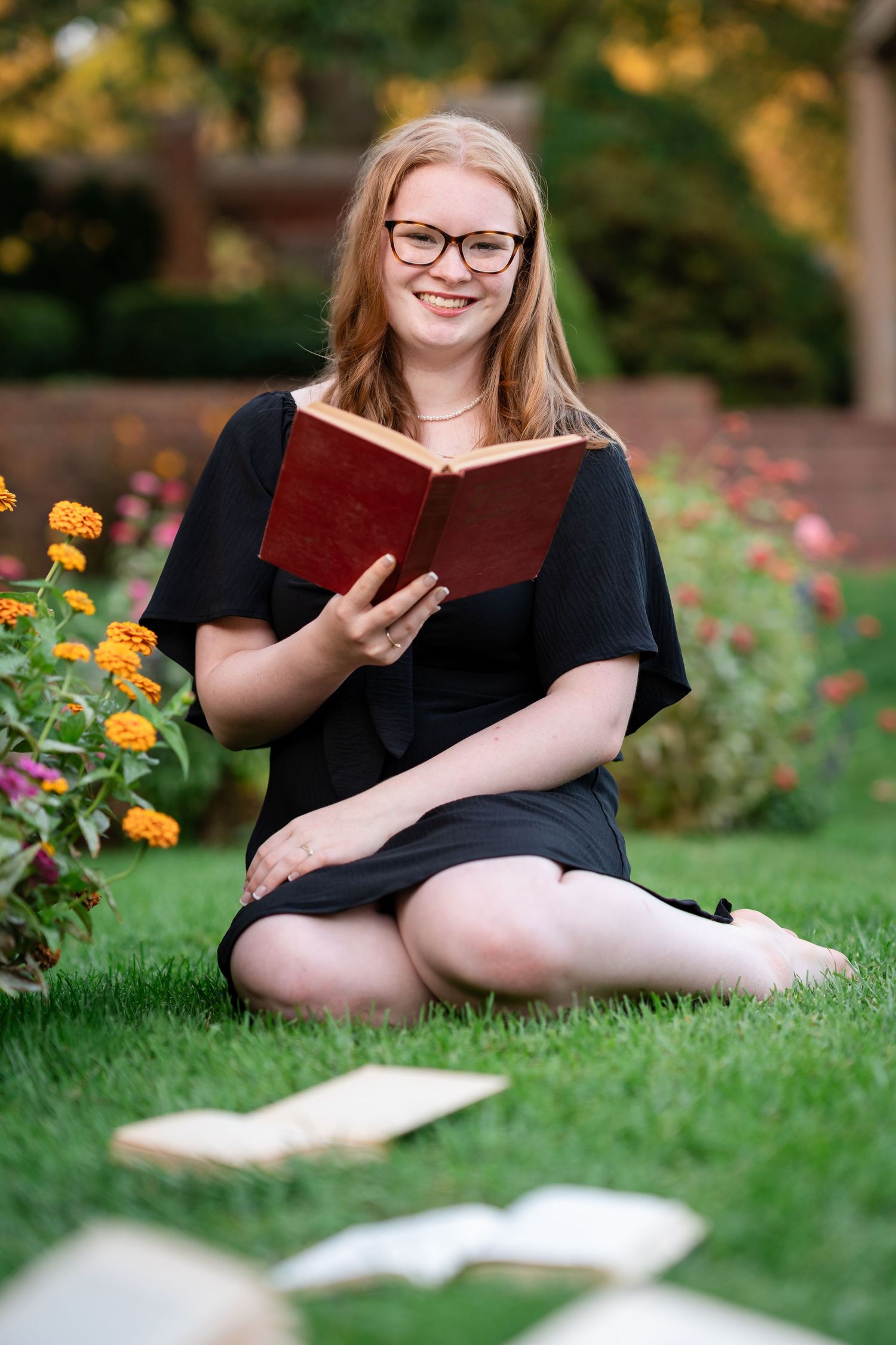 A person in black dress sits on grass reading a book in a garden with blooming flowers on a sunny day.