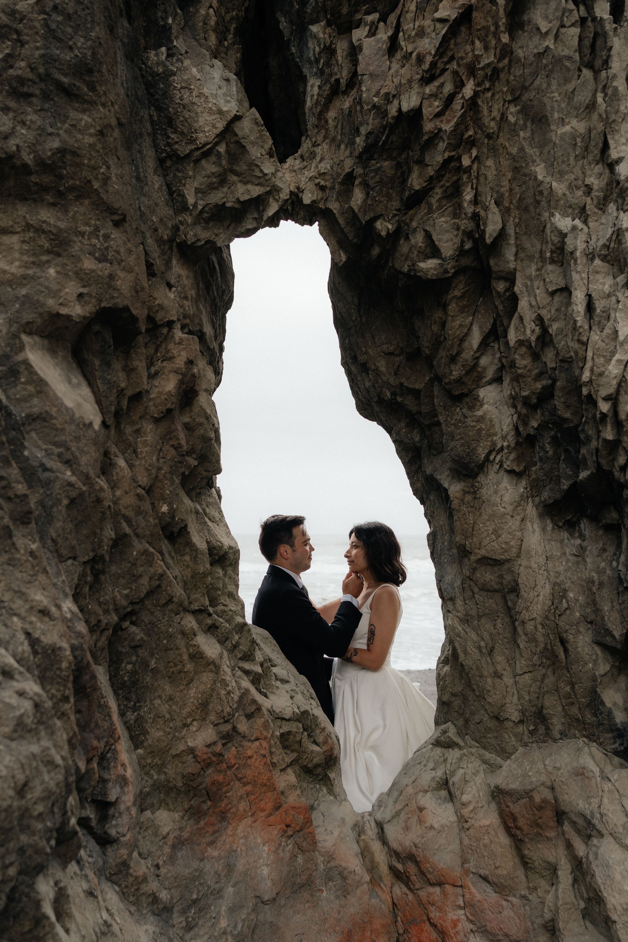 Couple in wedding attire embracing within a dramatic rocky arch formation with ocean views behind them.