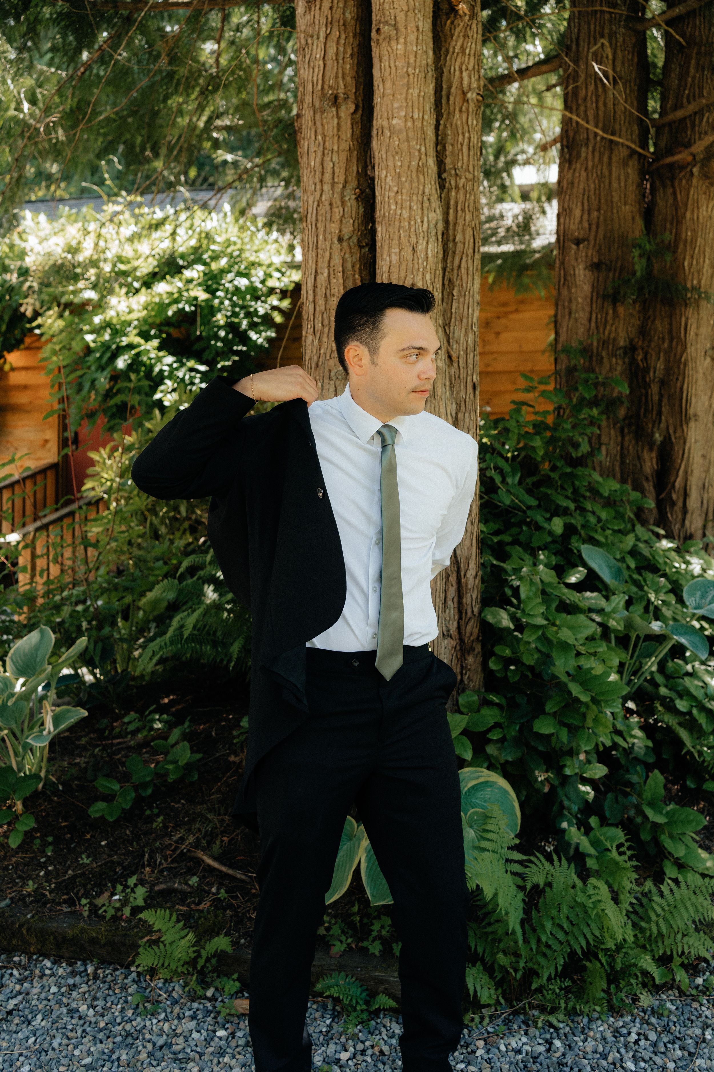 Groom in black suit and olive tie posing with jacket over shoulder in lush garden setting.