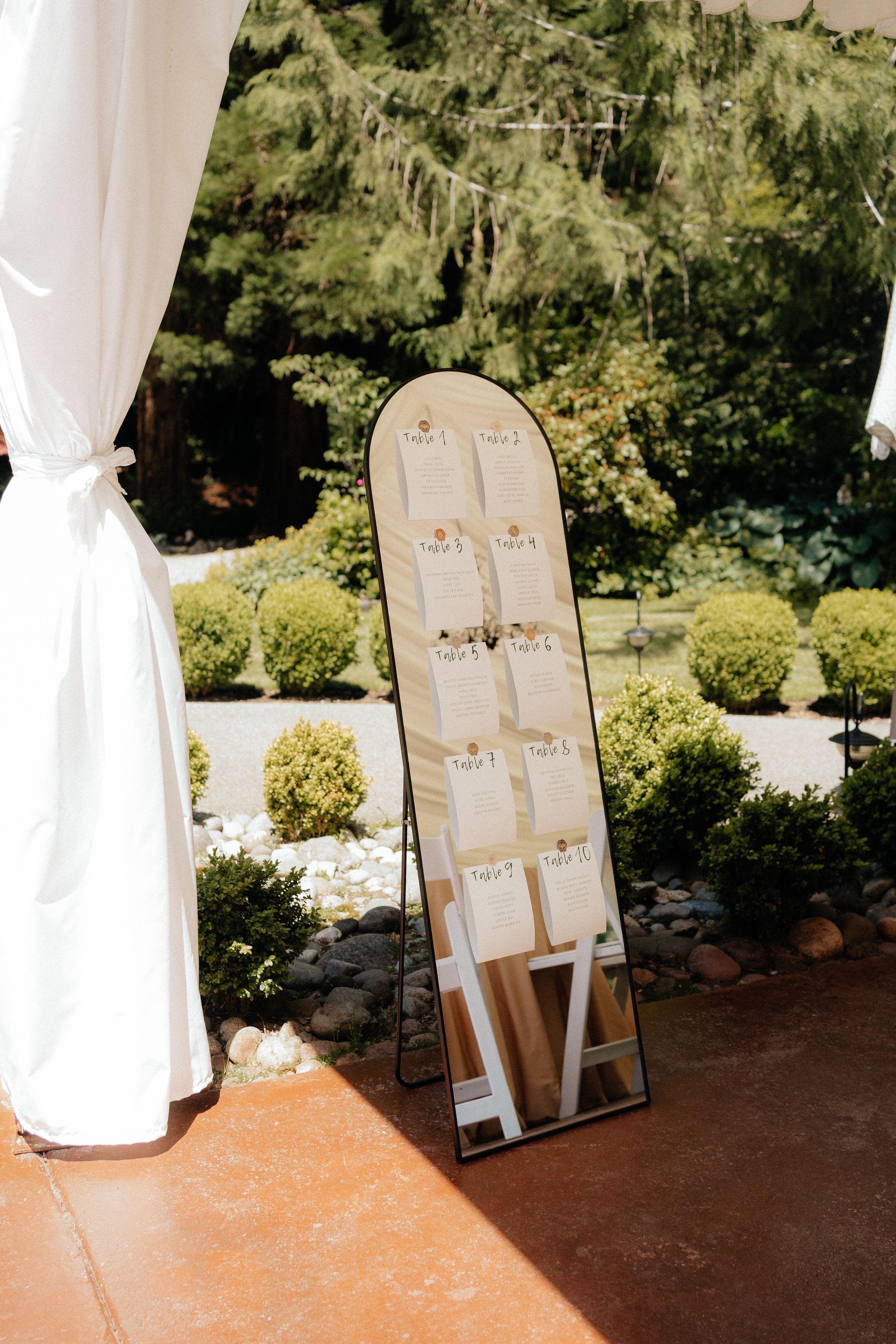 Elegant wedding seating chart displayed on arched mirror outdoors with white draping and greenery.