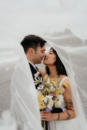 Bride and groom share a kiss under a flowing veil in the rain, bride holding a colorful bouquet.