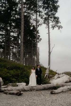Wedding couple standing on driftwood-covered beach surrounded by tall evergreen trees on overcast day.