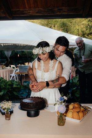 Bride and groom cutting chocolate wedding cake together at outdoor reception table with flowers.