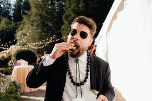 Man in black suit and sunglasses eating appetizer at outdoor wedding reception with string lights.