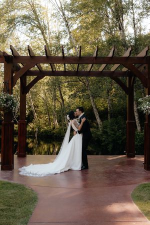Bride and groom share first kiss under wooden pergola with lush green trees and pond backdrop.