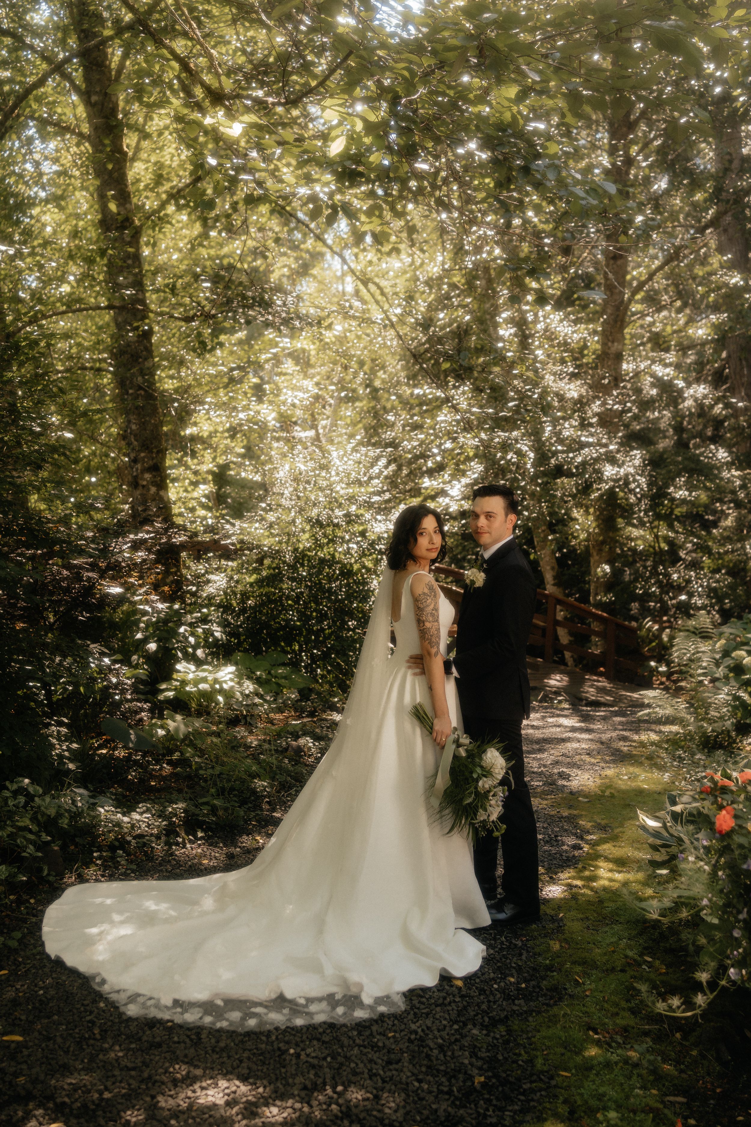 Bride and groom stand together on a garden path surrounded by lush green trees in a romantic outdoor wedding portrait.