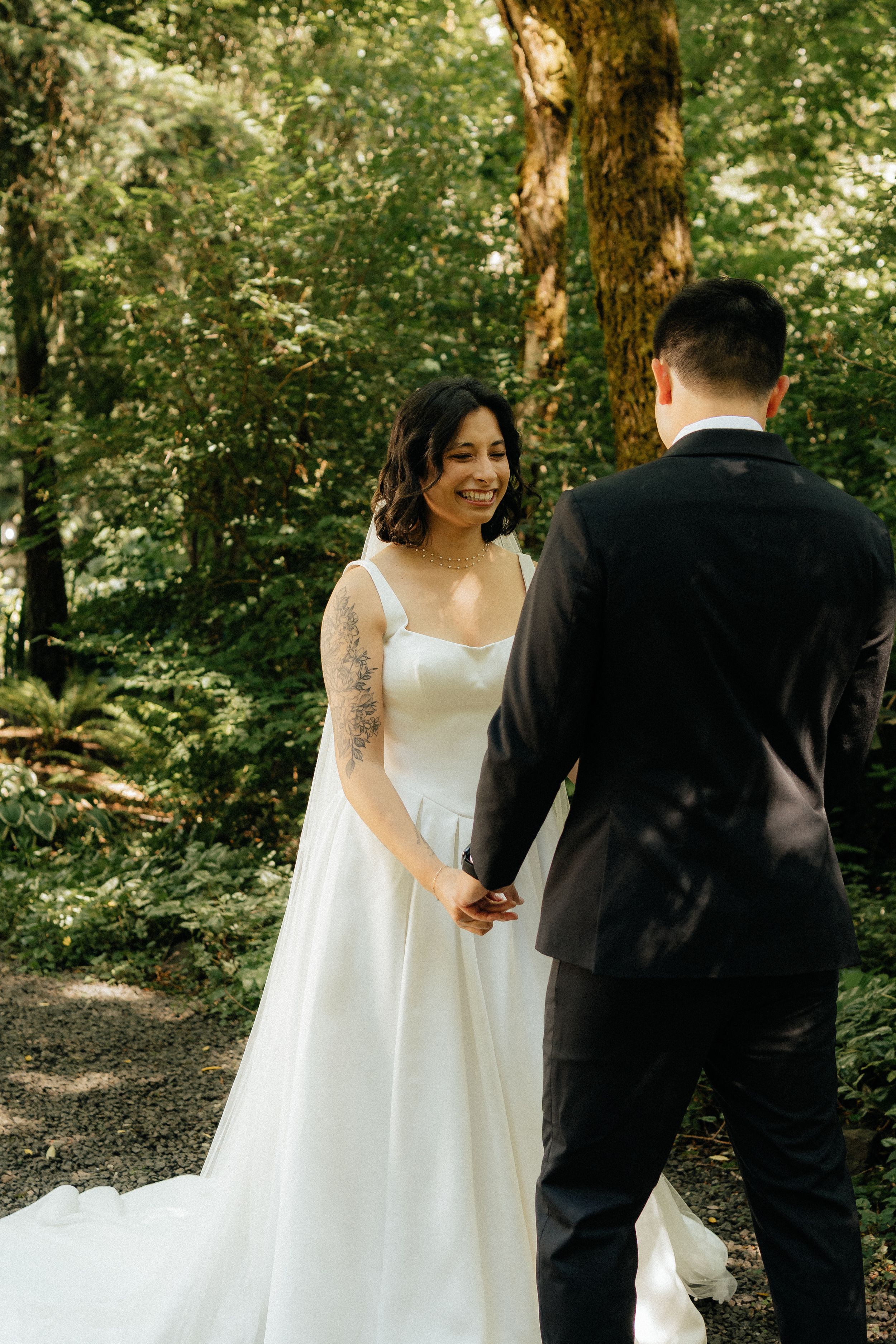 Smiling bride in white gown holds hands with groom in black suit during forest wedding.