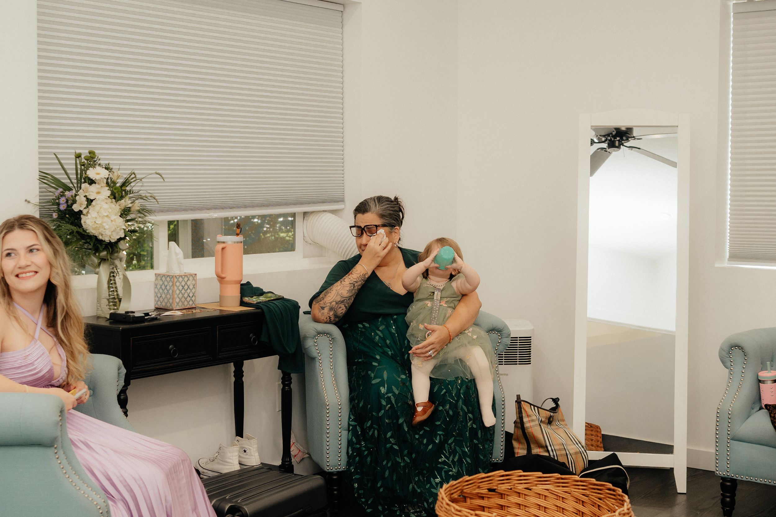 Woman in green dress holds baby while another woman in pink dress relaxes in bright room.