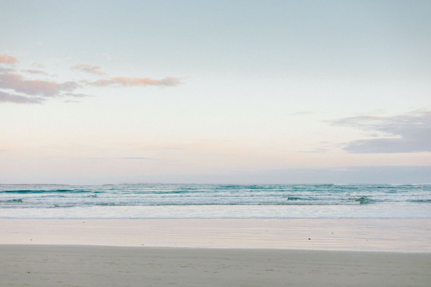A scenic coastal landscape featuring waves crashing on the beach at sunset with pink and purple clouds in the sky.