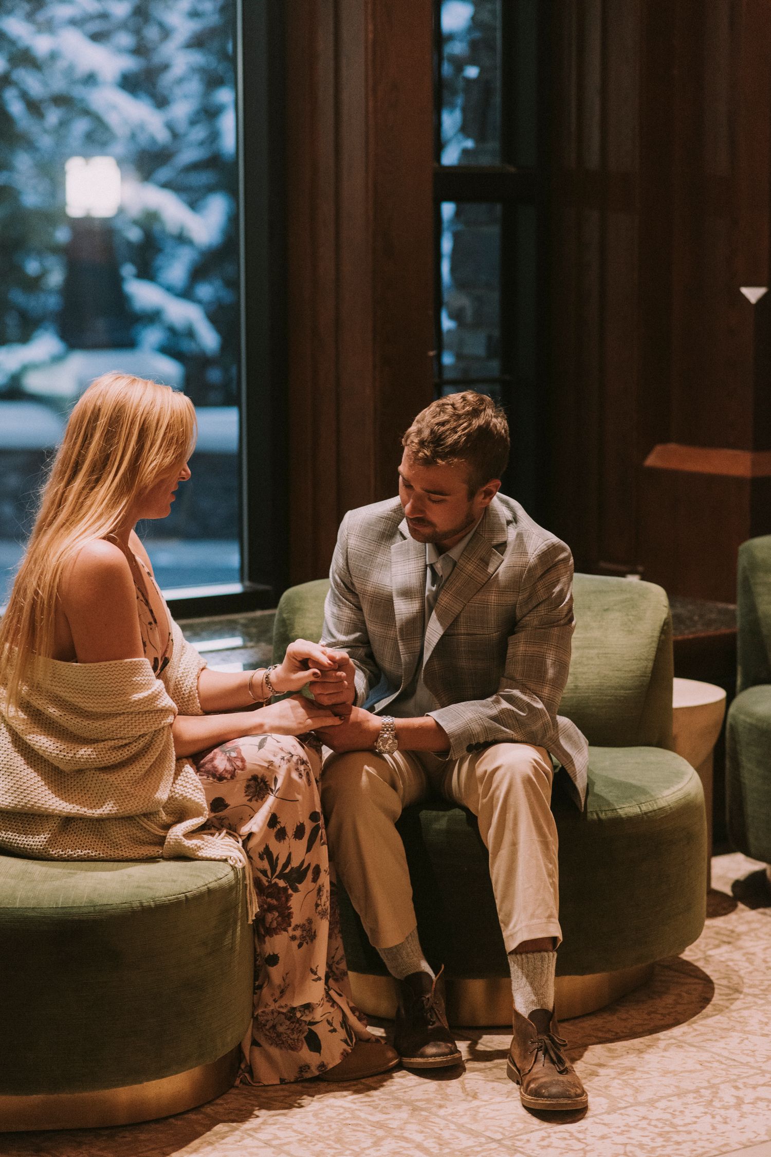 A couple sits together on green armchairs having an intimate conversation in a cozy indoor setting while looking at the engagement ring.