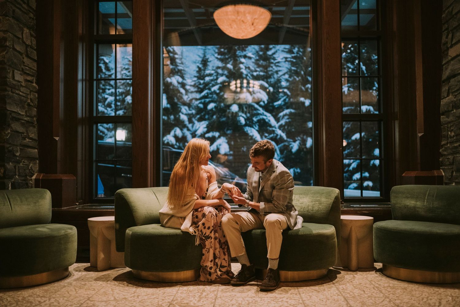 Couple sitting in cozy lodge chairs by large windows in Banff Spring Hotel overlooking snowy forest landscape during golden hour.