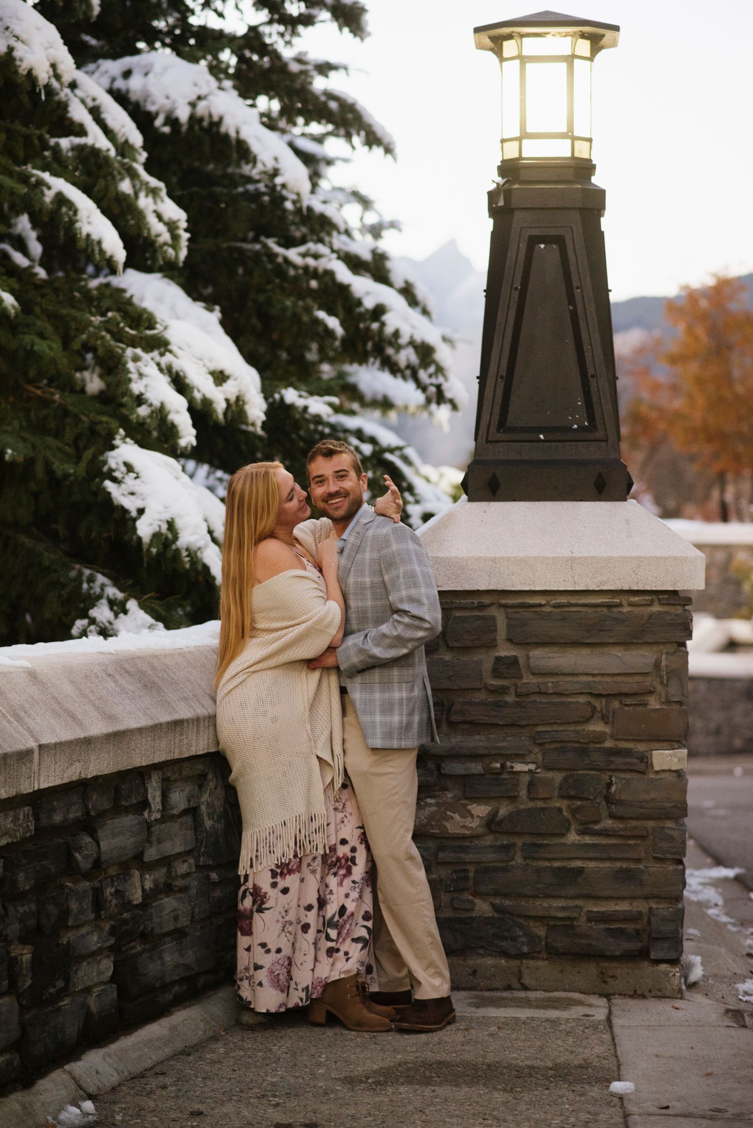 A couple shares a romantic moment on a stone wall with snow-covered evergreen trees in the background of Fairmont Banff Spring Hotel.