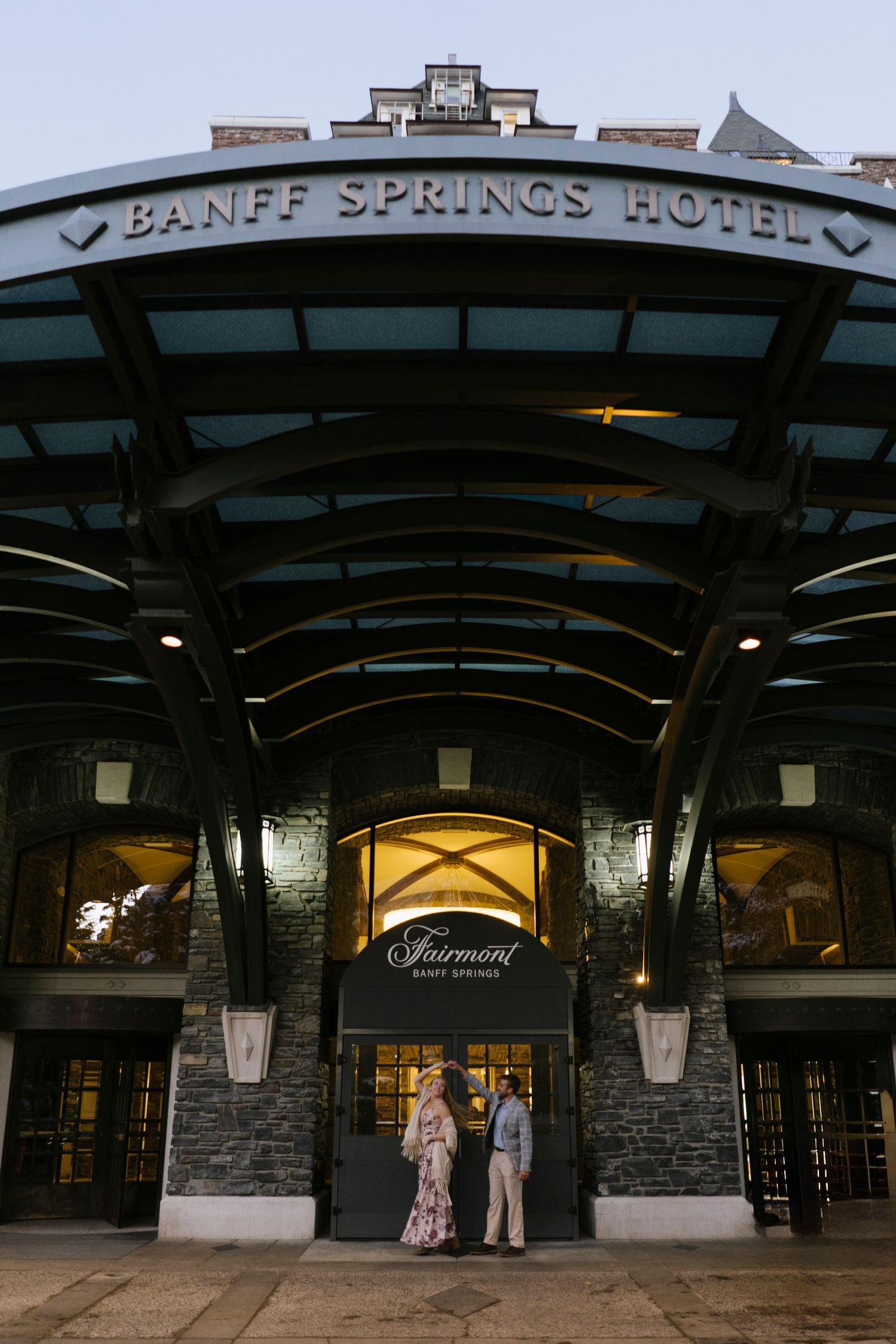 Illuminated entrance of the historic Banff Springs Hotel at dusk with stone facade and curved archway entrance.