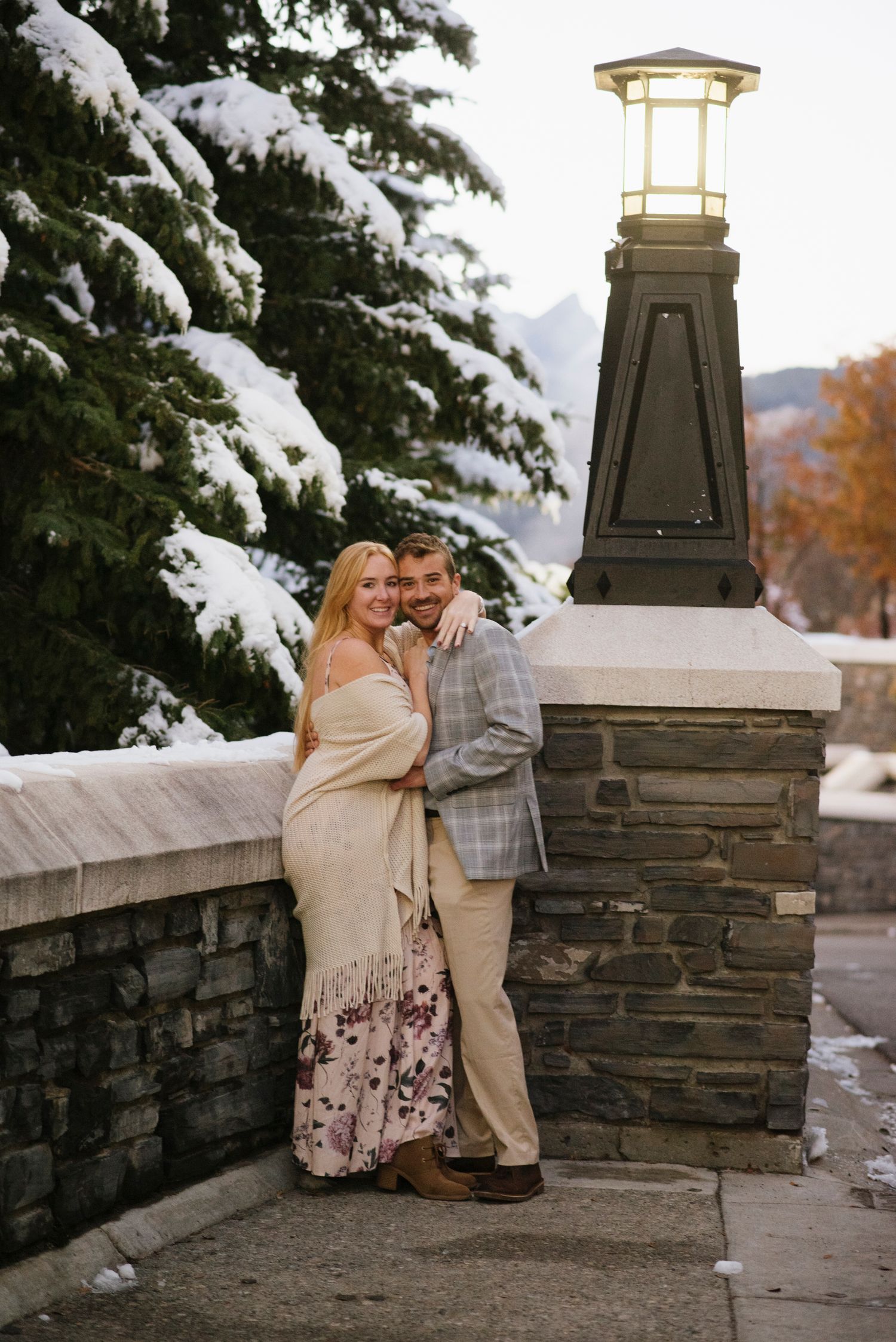A couple shares a romantic moment on a stone wall with snow-covered evergreen trees in the background of Fairmont Banff Spring Hotel.