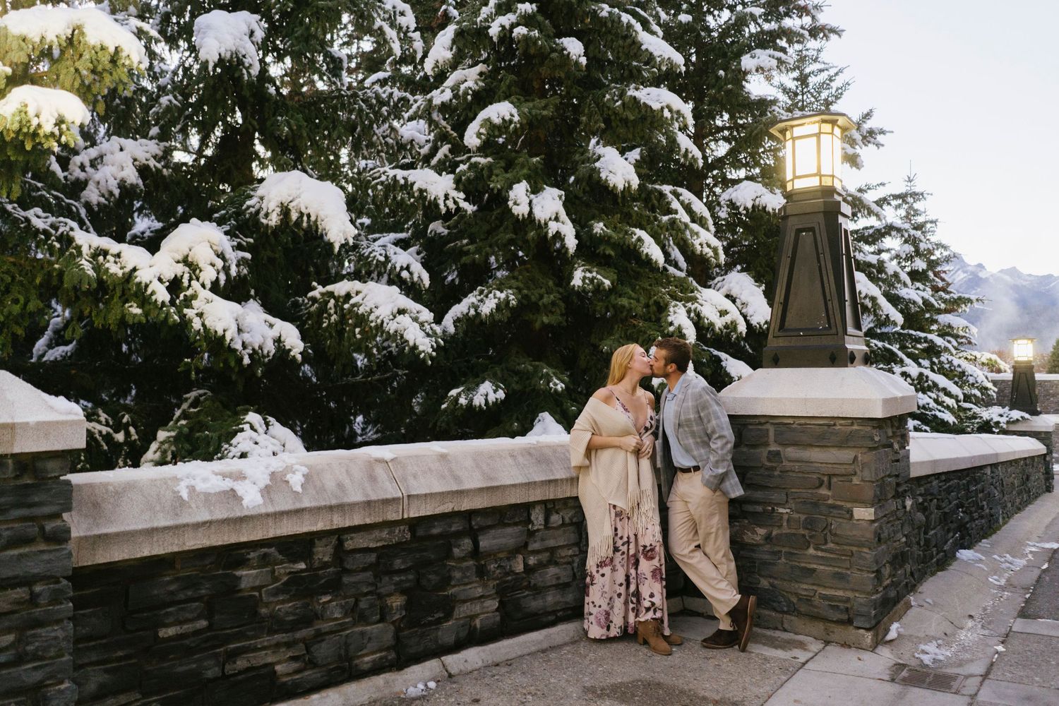 A couple shares a romantic moment on a stone wall with snow-covered evergreen trees in the background of Fairmont Banff Spring Hotel.