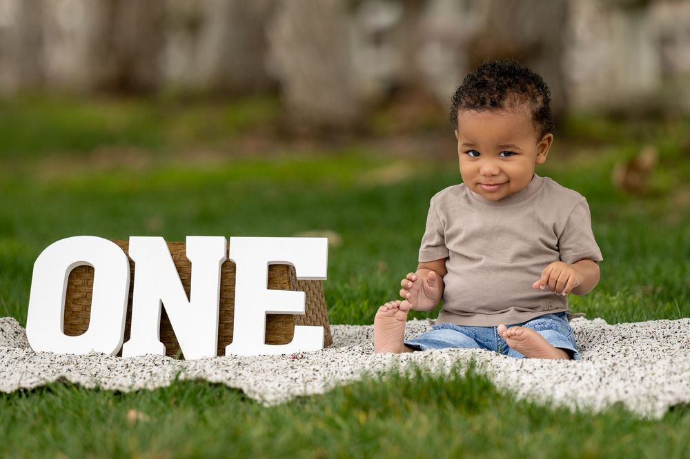A toddler sits on white fabric next to large decorative 'ONE' letters on grass.