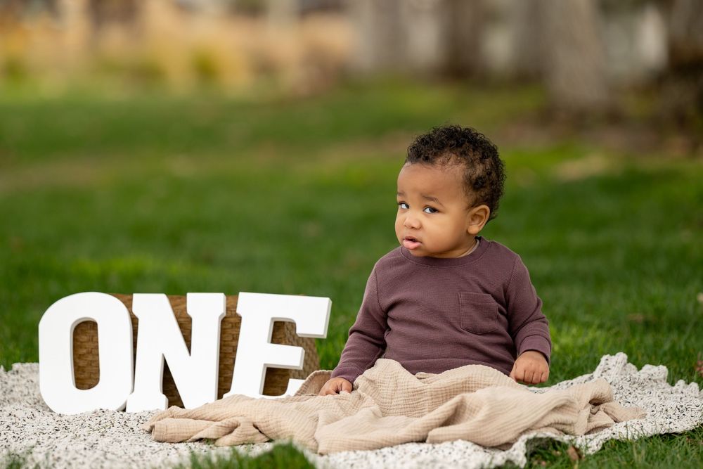 A small child in a brown sweater sits beside decorative 'ONE' letters on white fabric.