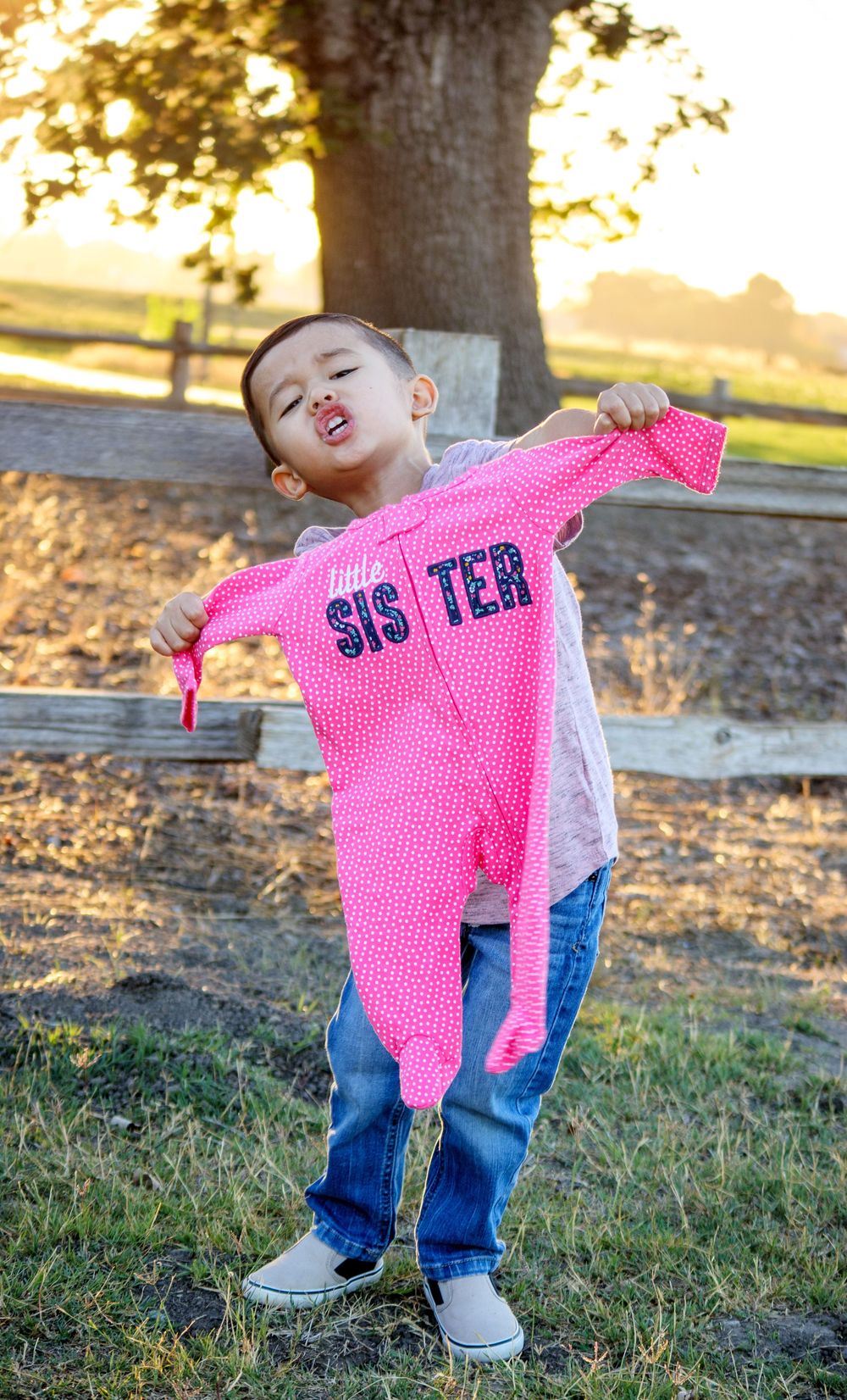 Child holding pink shirt with 'Sister' text while playing outdoors at sunset.
