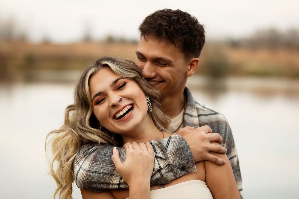 A couple shares a romantic moment laughing and embracing by a lake on an overcast day.