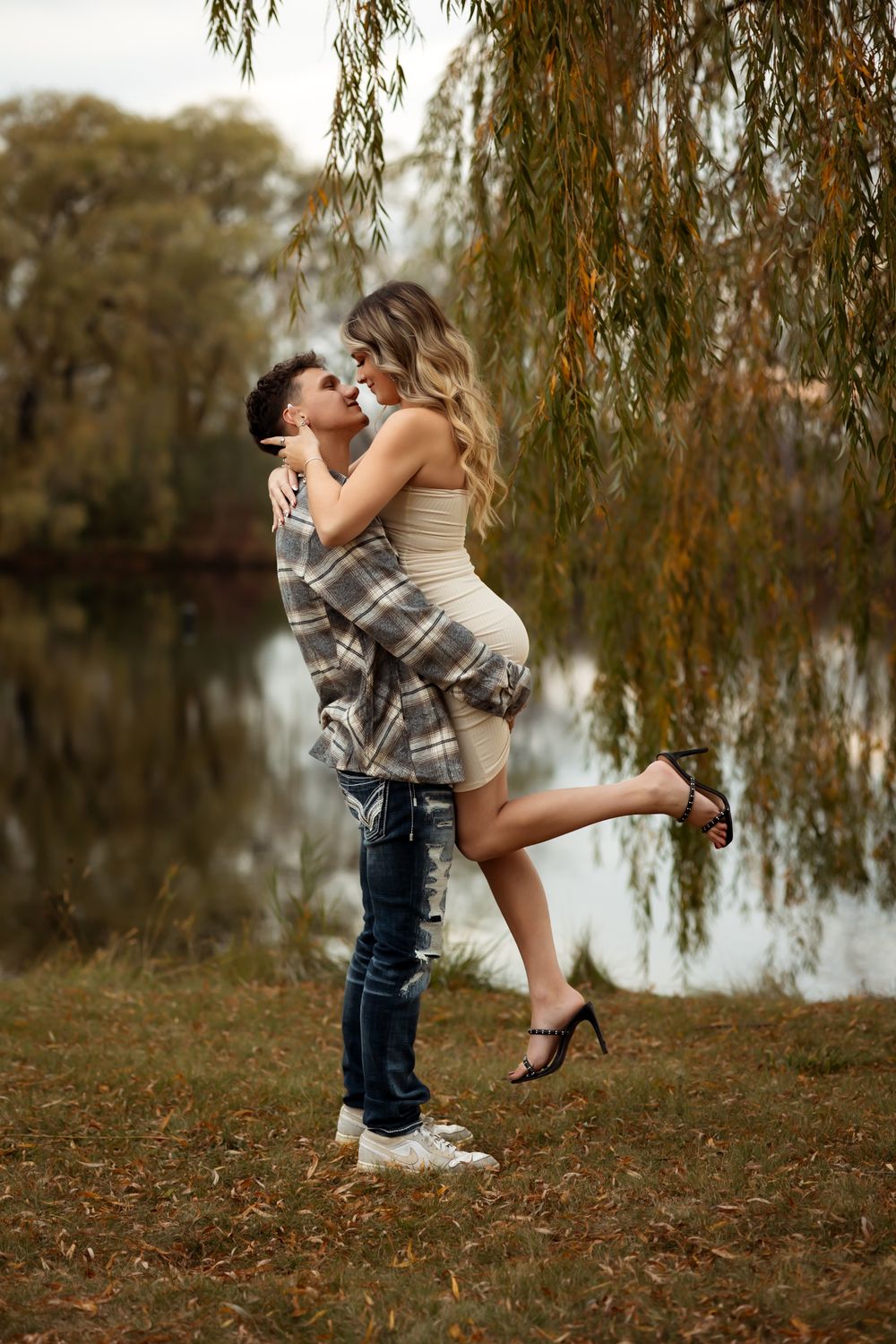 A romantic couple embraces under a willow tree by a lake in autumn as fallen leaves scatter the ground.