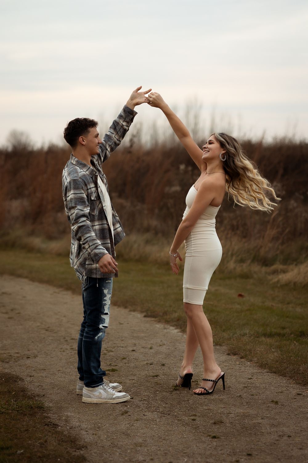 A couple in contrasting outfits, white dress and plaid shirt, dance playfully on a dirt path at sunset.