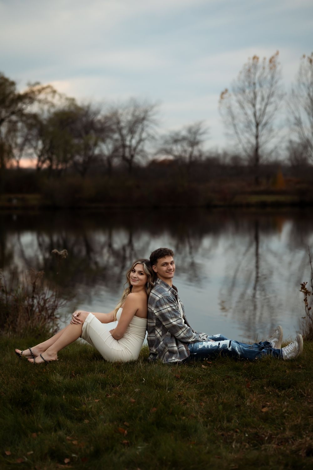 Couple relaxing on grassy hill beside a serene lake during autumn evening.