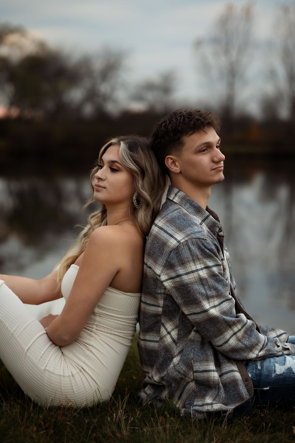 Couple in casual attire sitting back to back by a serene lake during golden hour.