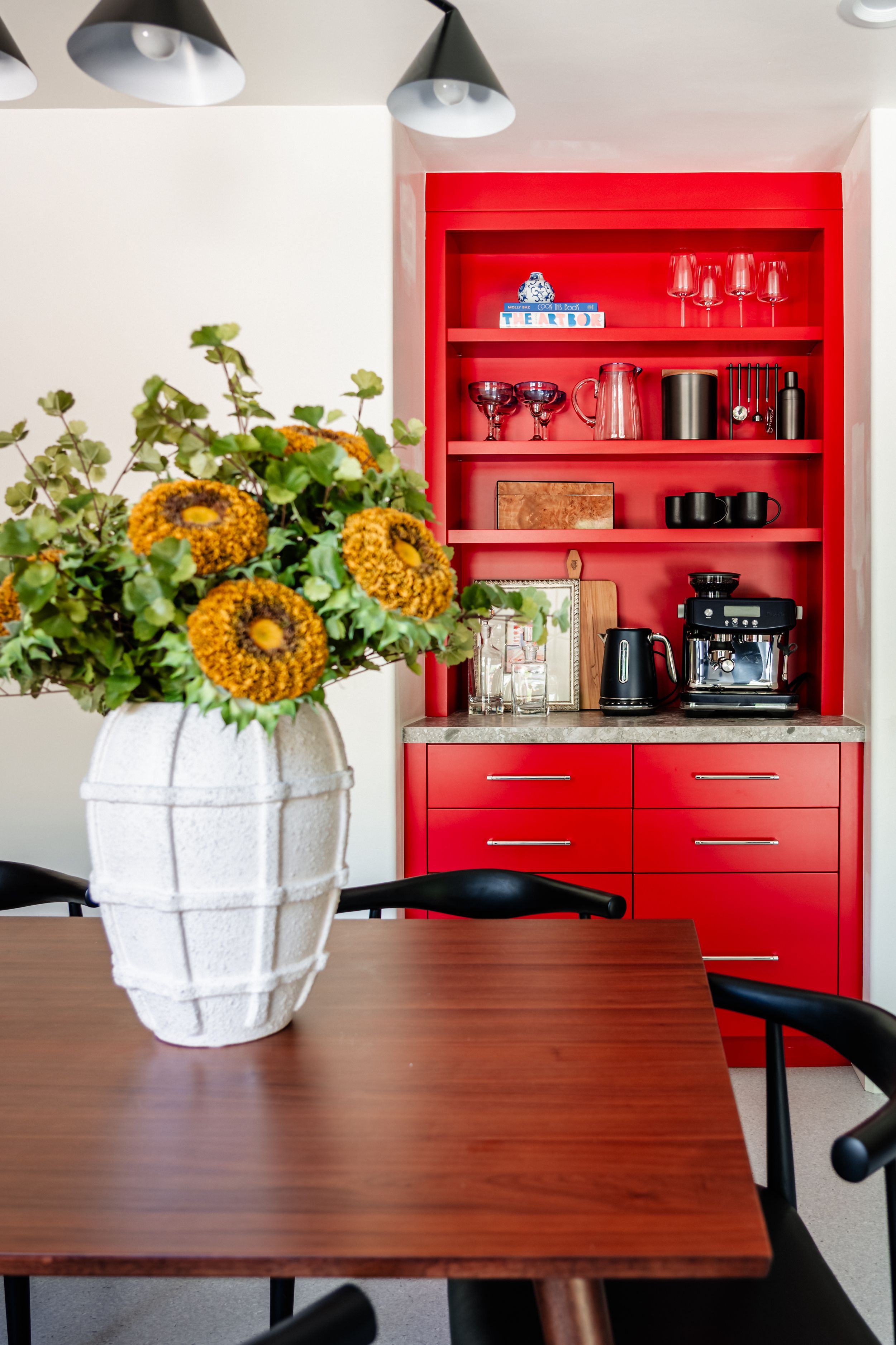 A modern dining room with orange flowers in a white vase and a bold red built-in cabinet with coffee station.