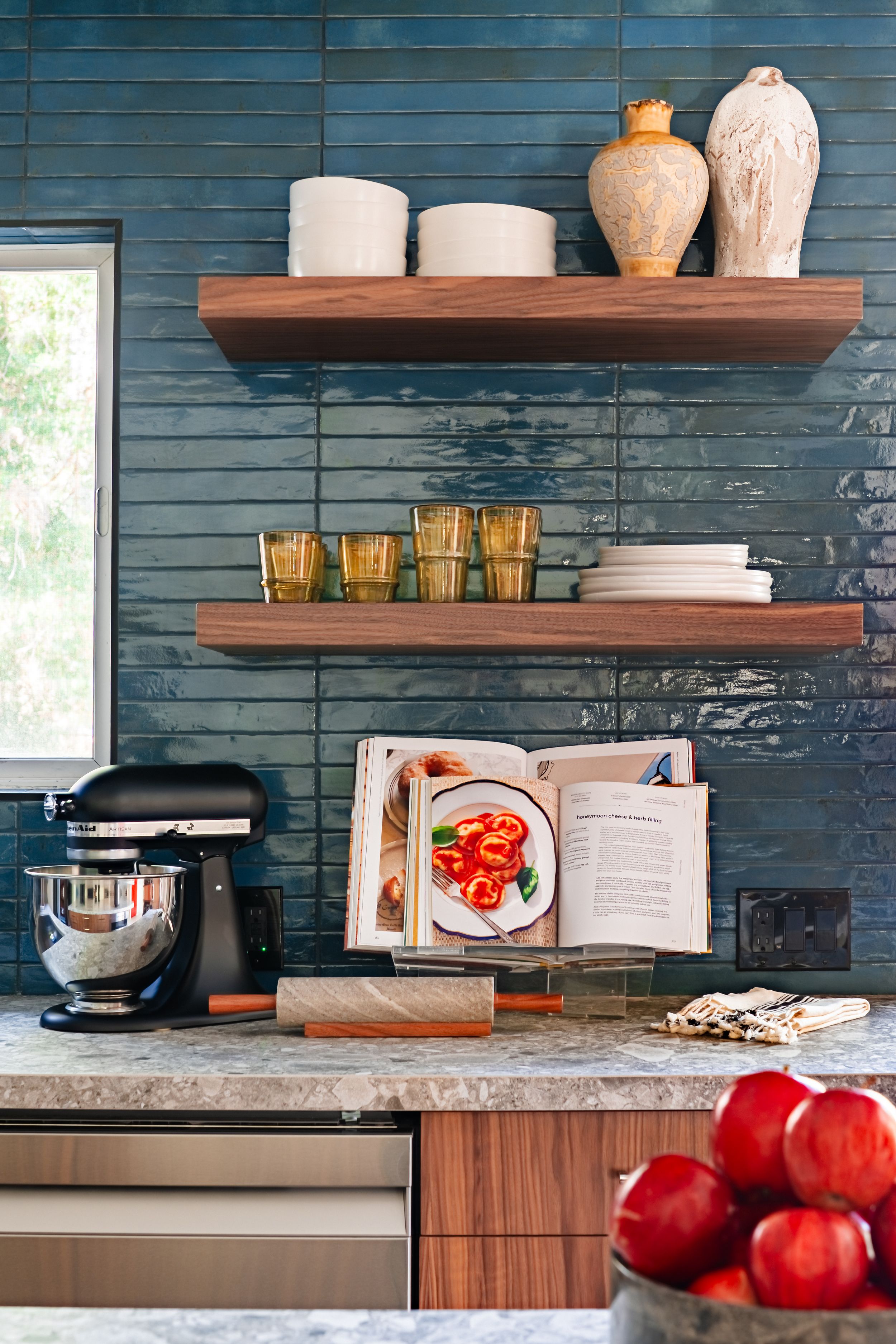 Modern kitchen interior with blue tile backsplash, wooden shelves, and red apples on marble countertop.