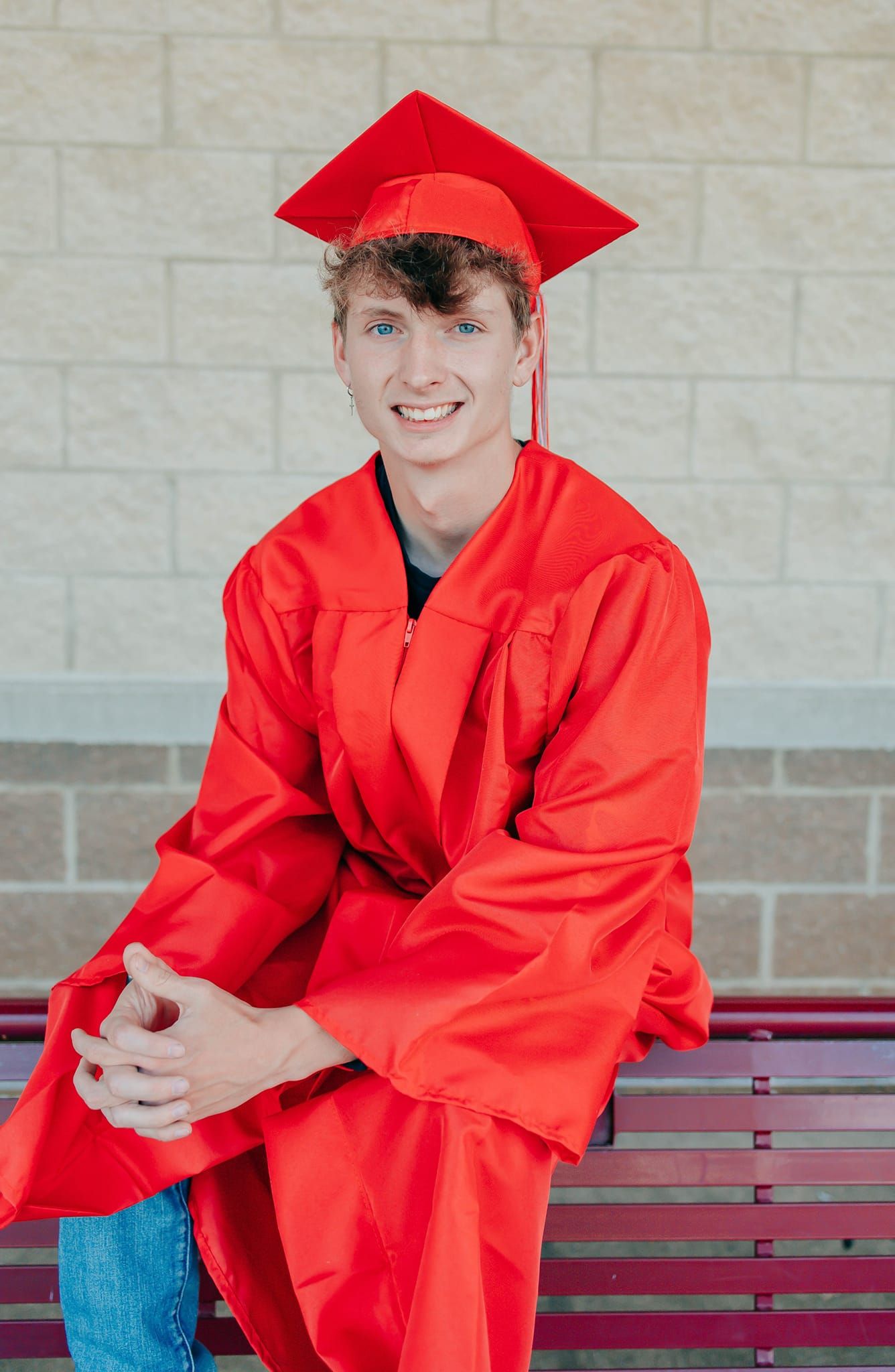 Graduate in bright red cap and gown sits casually on a wooden bench against a stone wall.