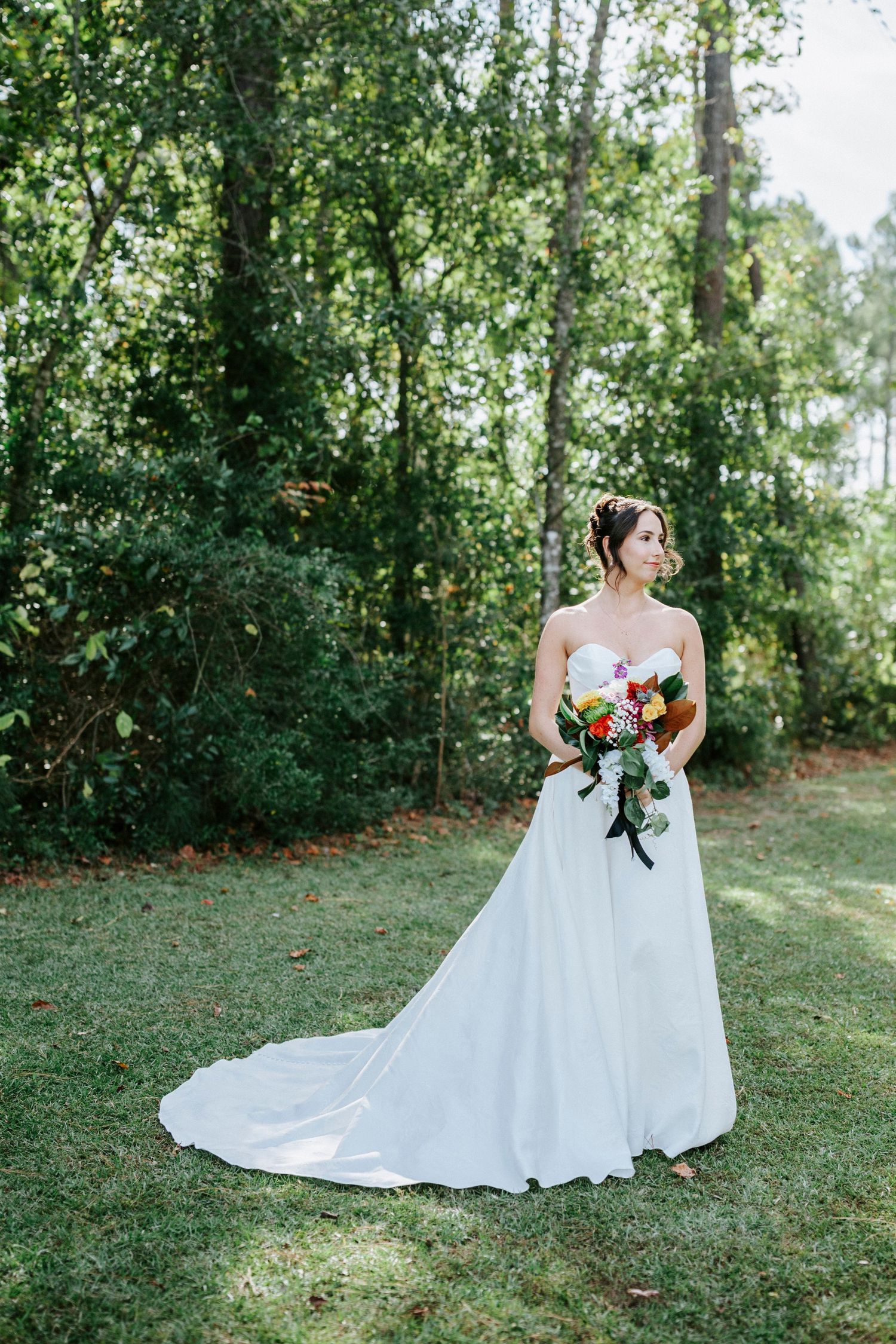 A bride in a flowing white wedding gown poses gracefully on a lush green lawn surrounded by trees.
