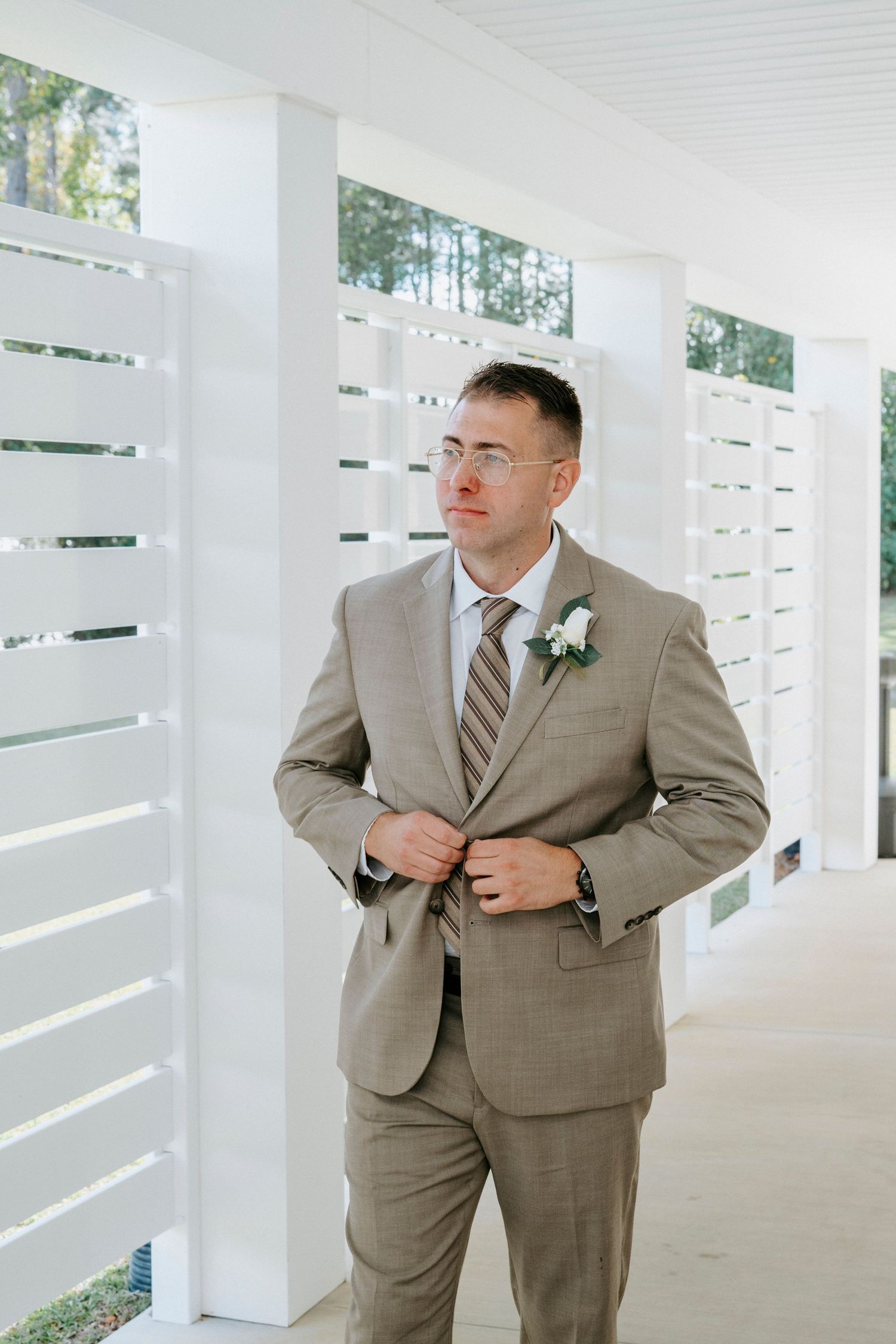 Someone in a tan suit and striped tie adjusts their jacket while standing in a white columned porch area.