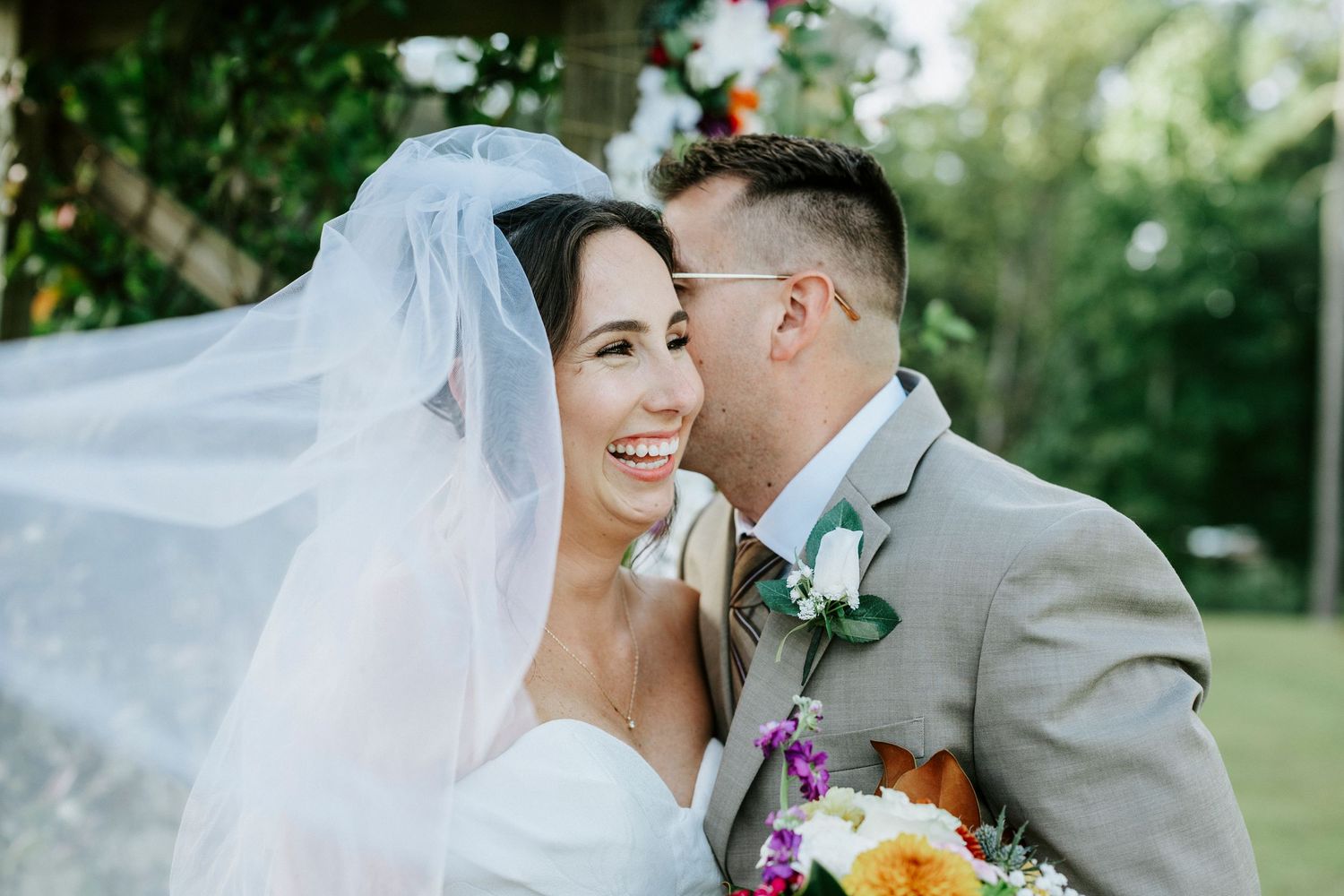 A couple shares intimate moments in their wedding attire while surrounded by lush greenery in an outdoor setting.