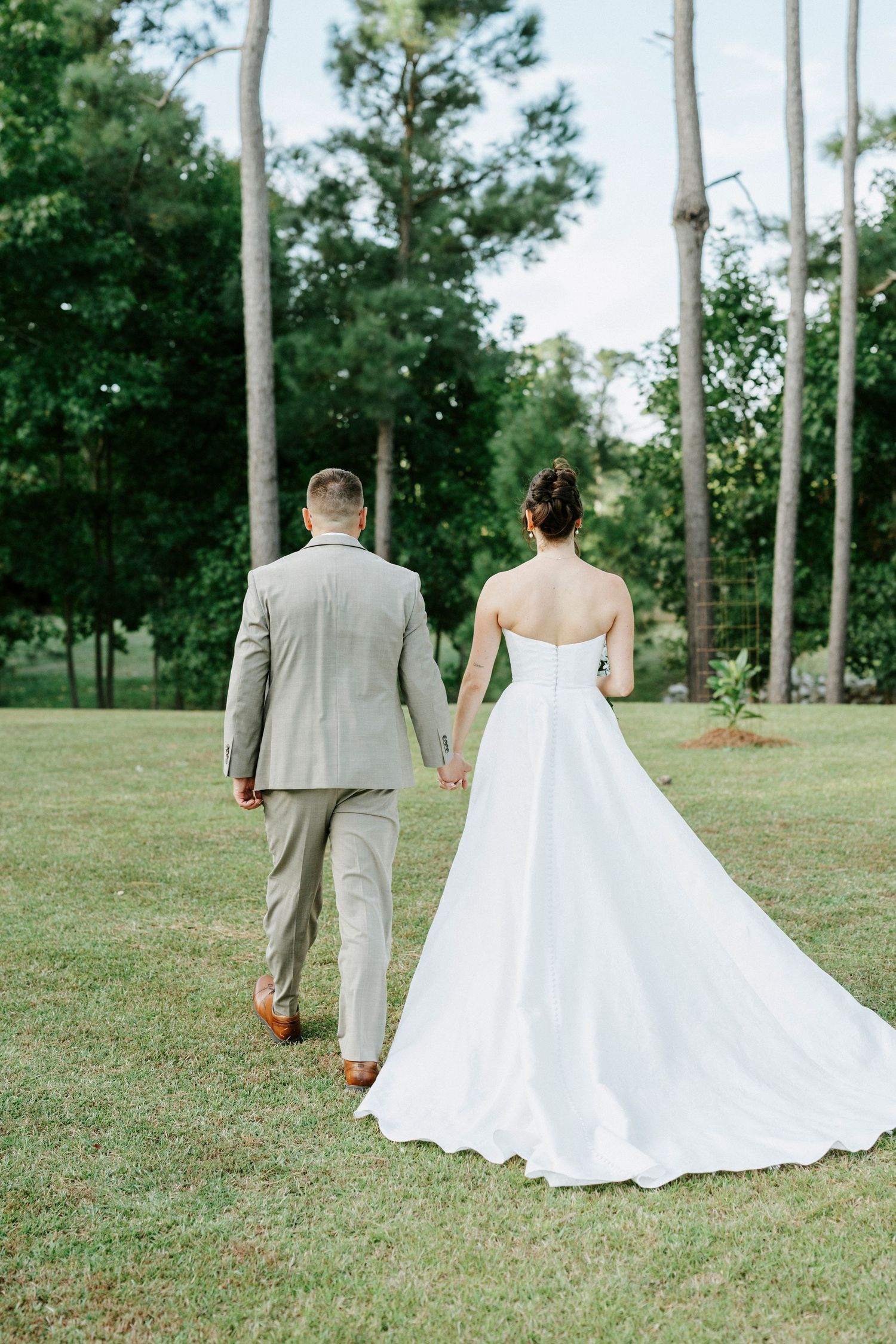A couple walks through a scenic tree-lined path, with the bride in white and groom in grey suit.