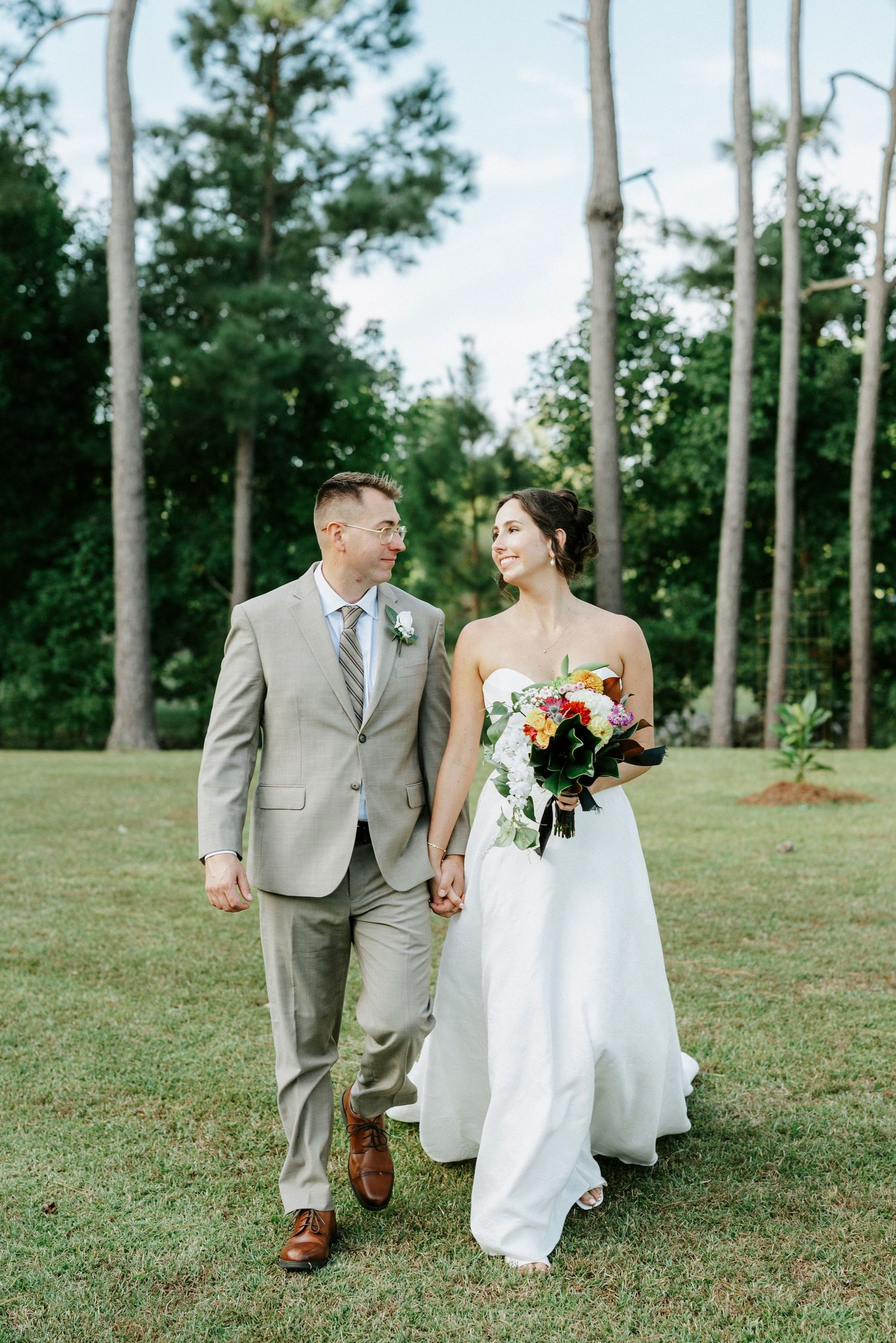 A couple in wedding attire walks hand in hand across a grassy lawn dotted with tall pine trees in the background.