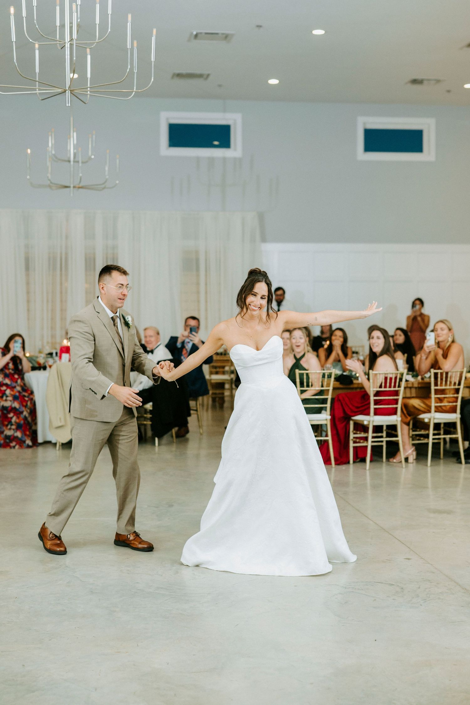 Couple performs their first dance at a modern wedding reception venue with white curtains and chandeliers.