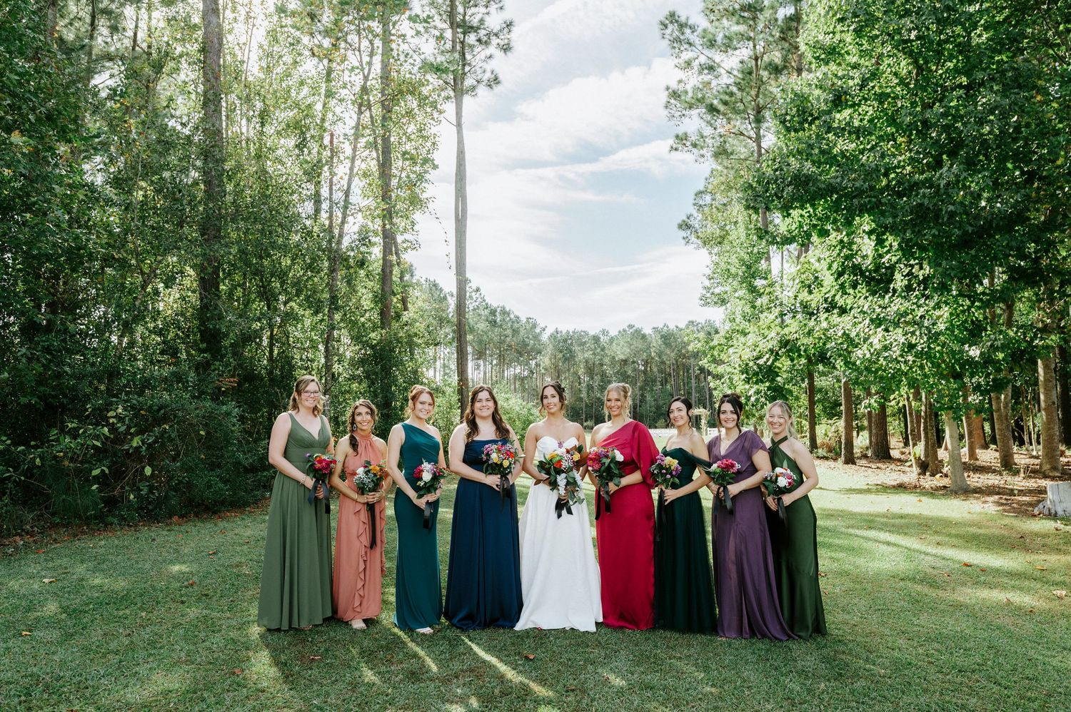 Wedding party stands together in a line on a grassy lawn surrounded by tall trees and natural scenery.