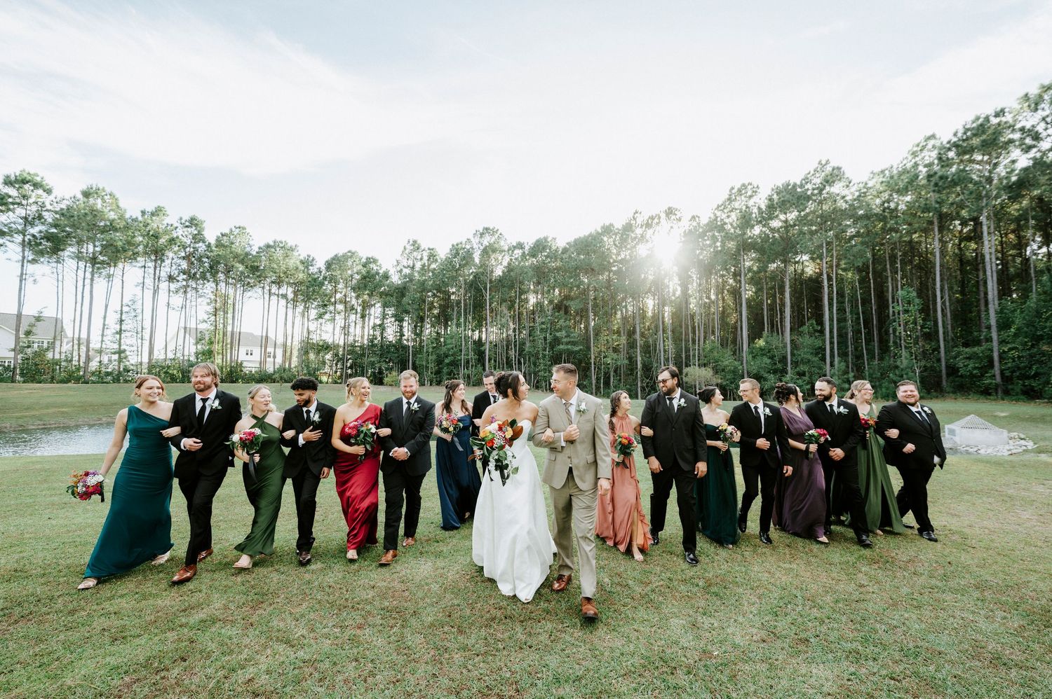 Wedding party poses together in a scenic outdoor setting with trees and sunlight in the background.