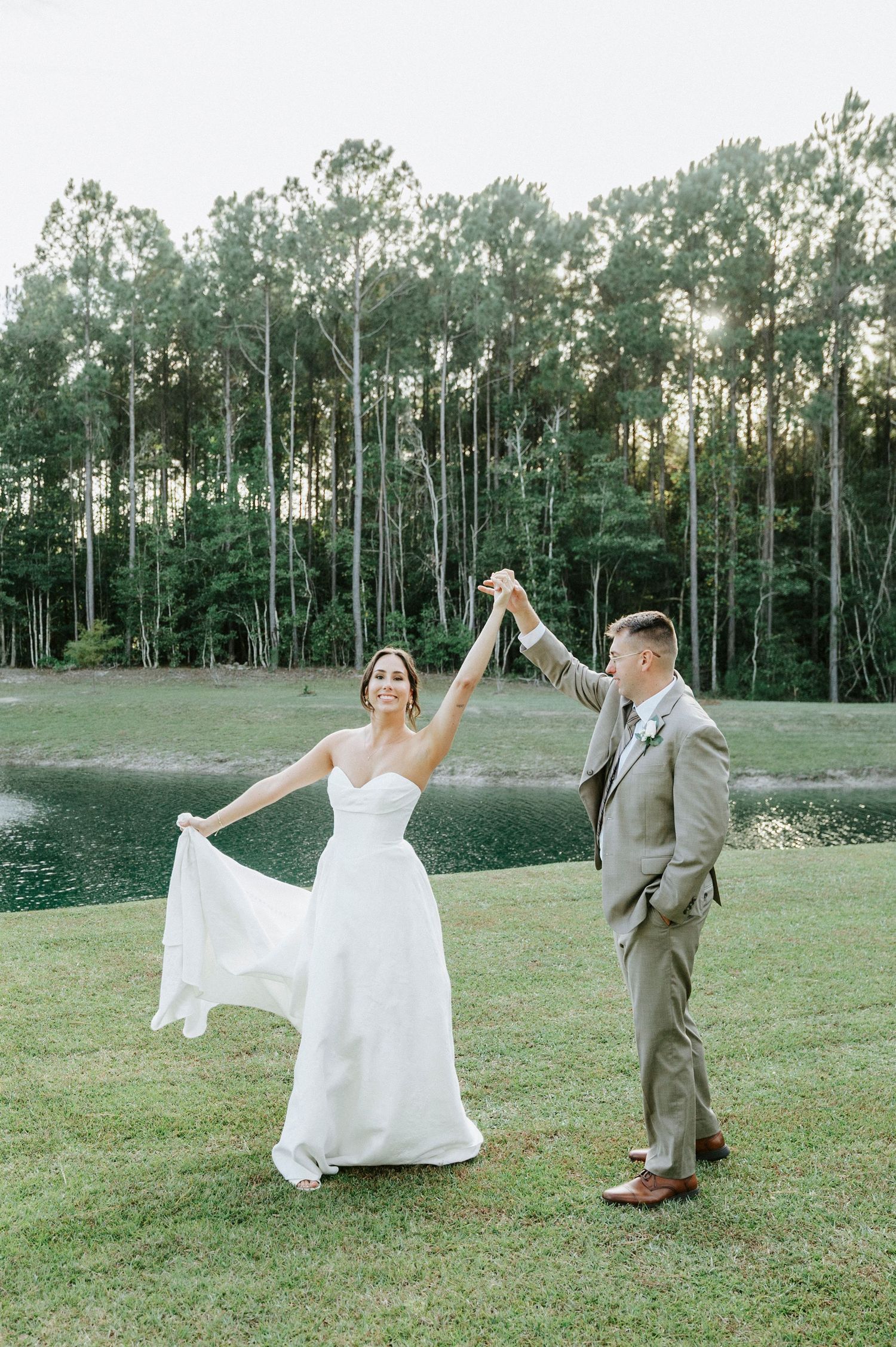A couple in wedding attire dances and twirls together on a grassy field with tall pine trees in the background.