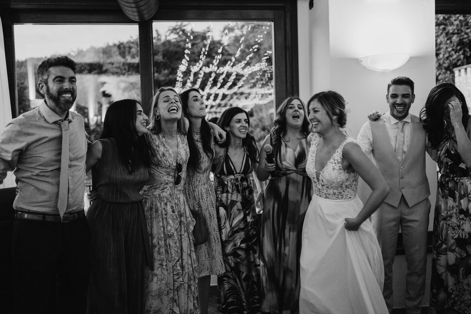 Black and white image of bride and guests laughing together joyfully at an indoor wedding celebration.