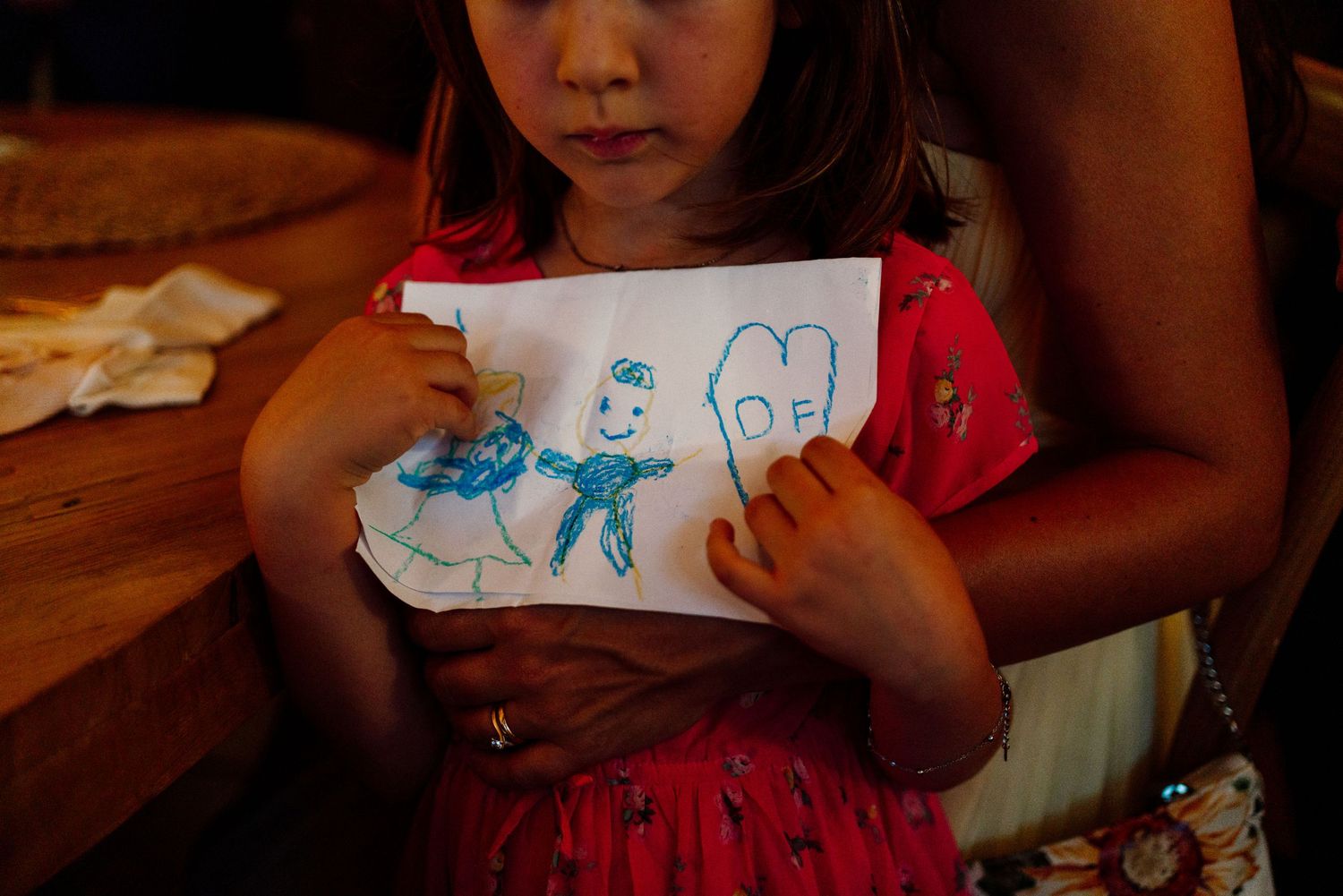 Young girl in red dress proudly holds a crayon drawing at a wedding reception dinner table.