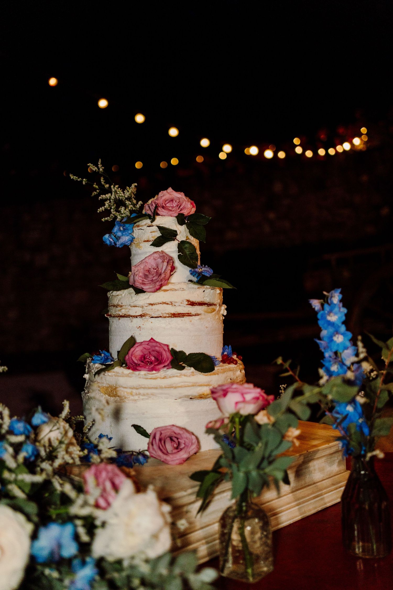 Elegant three-tier naked wedding cake adorned with pink and blue flowers under string lights at night.