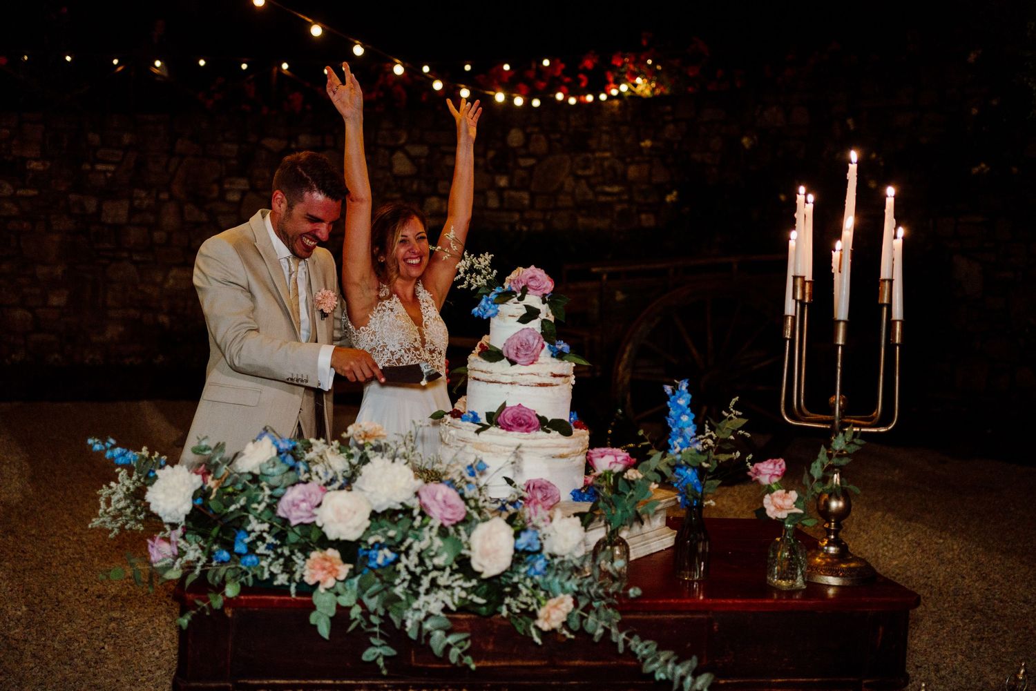 Excited couple raises arms while cutting their floral-decorated wedding cake by candlelight under string lights.