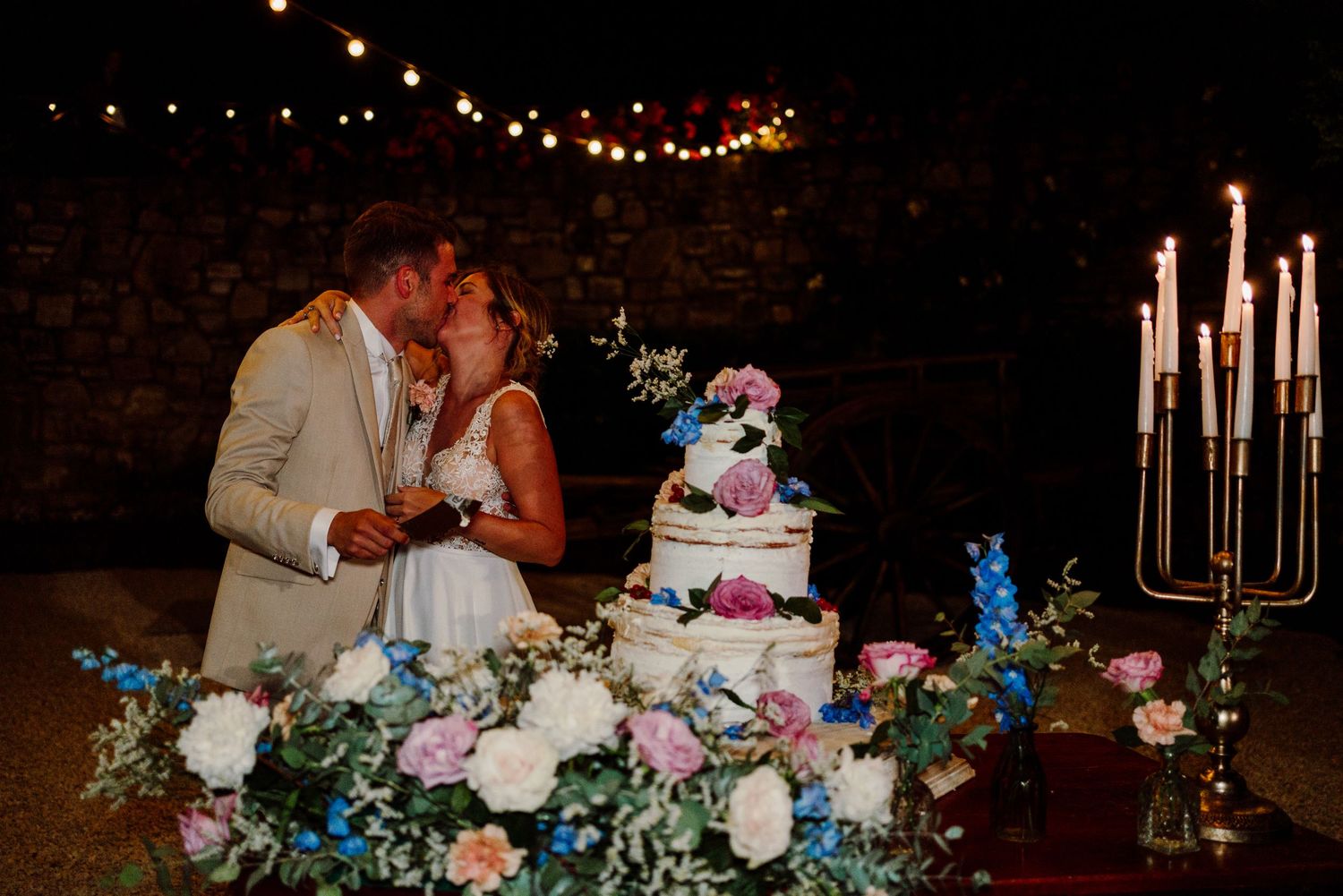Newlyweds share a tender kiss beside their decorated wedding cake with candles and colorful flowers at night.