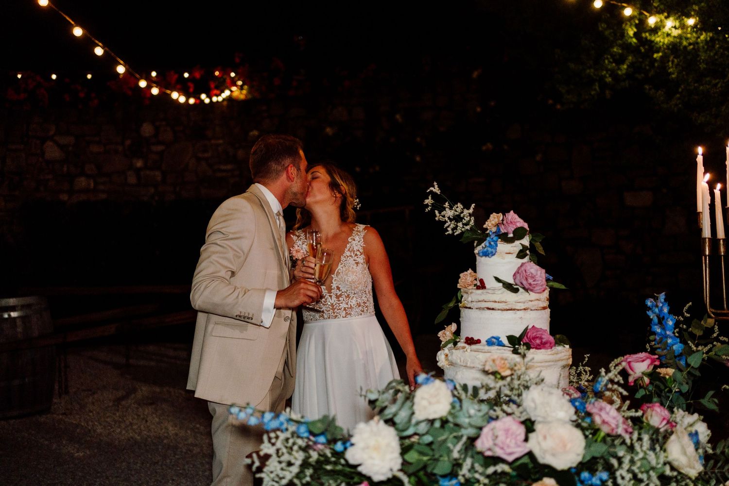 Couple shares a romantic kiss next to a floral wedding cake under warm string lights at night.