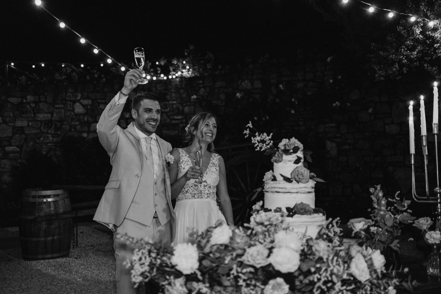 Black and white photo of bride and groom toasting with champagne beside wedding cake at outdoor night reception.